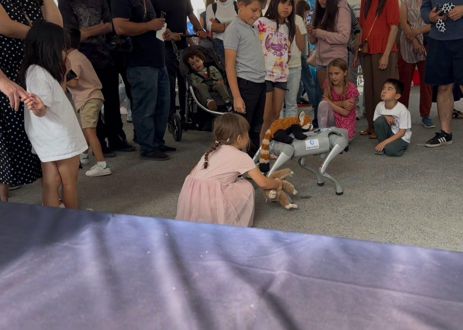 A child pets a Unitree Go2 robot dog wearing a plush costume while a crowd looks on